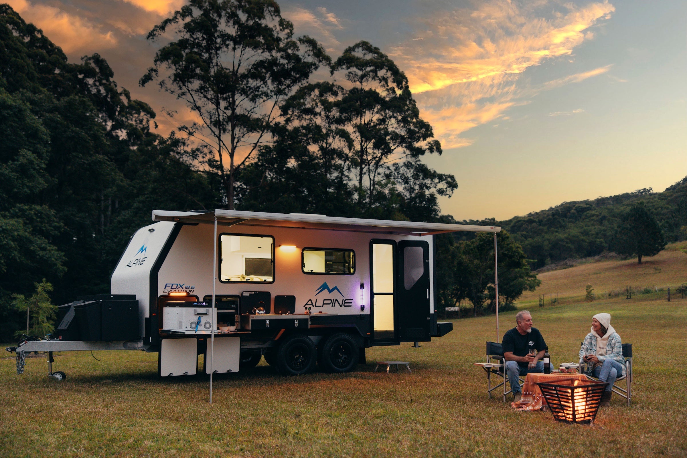 Two people sitting by a campfire in front of a caravan in a scenic outdoor setting.